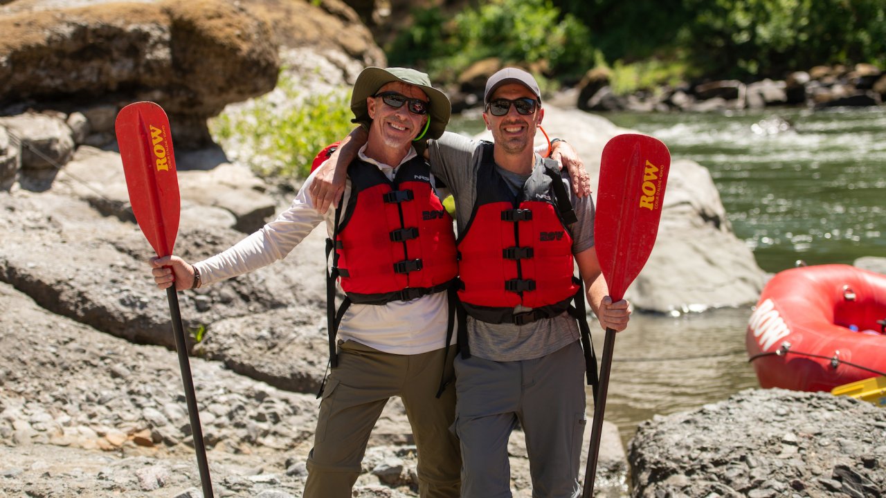 Two people wearing sunglasses, hats, and life jackets holding their paddles smile for a photo