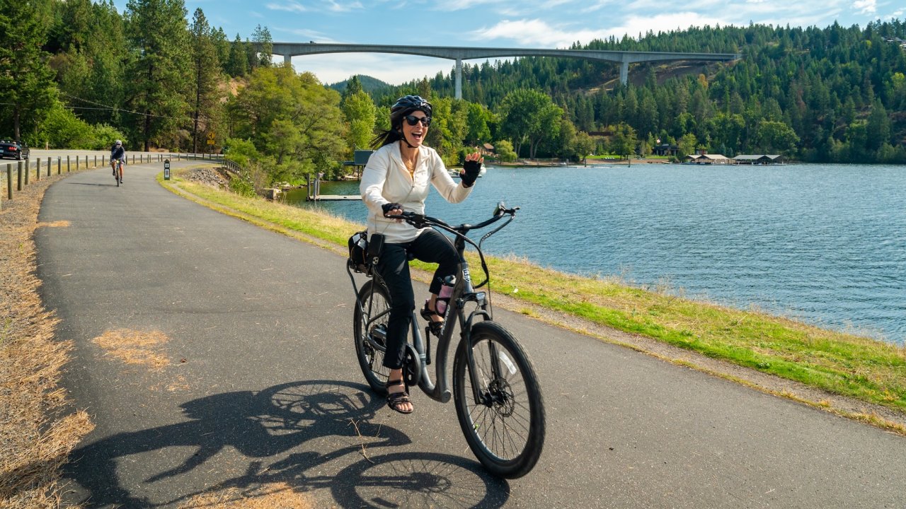 Smiling woman rides an e-bike along a paved trail by Lake Coeur d’Alene in North Idaho, a popular biking destination