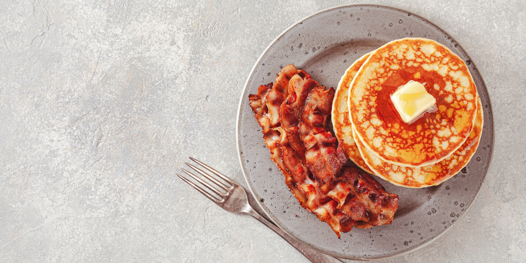 Top down view of pancakes with a slab of butter and crispy bacon on a grey plate with a fork on the side