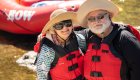 Smiling couple wearing life jackets and sun hats on the Clearwater River in Idaho, standing next to a red ROW Adventures rafting boat — enjoying a scenic Idaho rafting trip for all ages.
