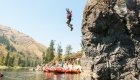 Person cliff jumping into a river near red rafts and a group onshore.