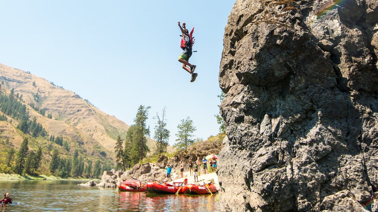 Person cliff jumping into a river near red rafts and a group onshore.