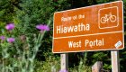 A brown trailhead sign reading 'Route of the Hiawatha – West Portal' stands beside blooming wildflowers and a lush green forest.
