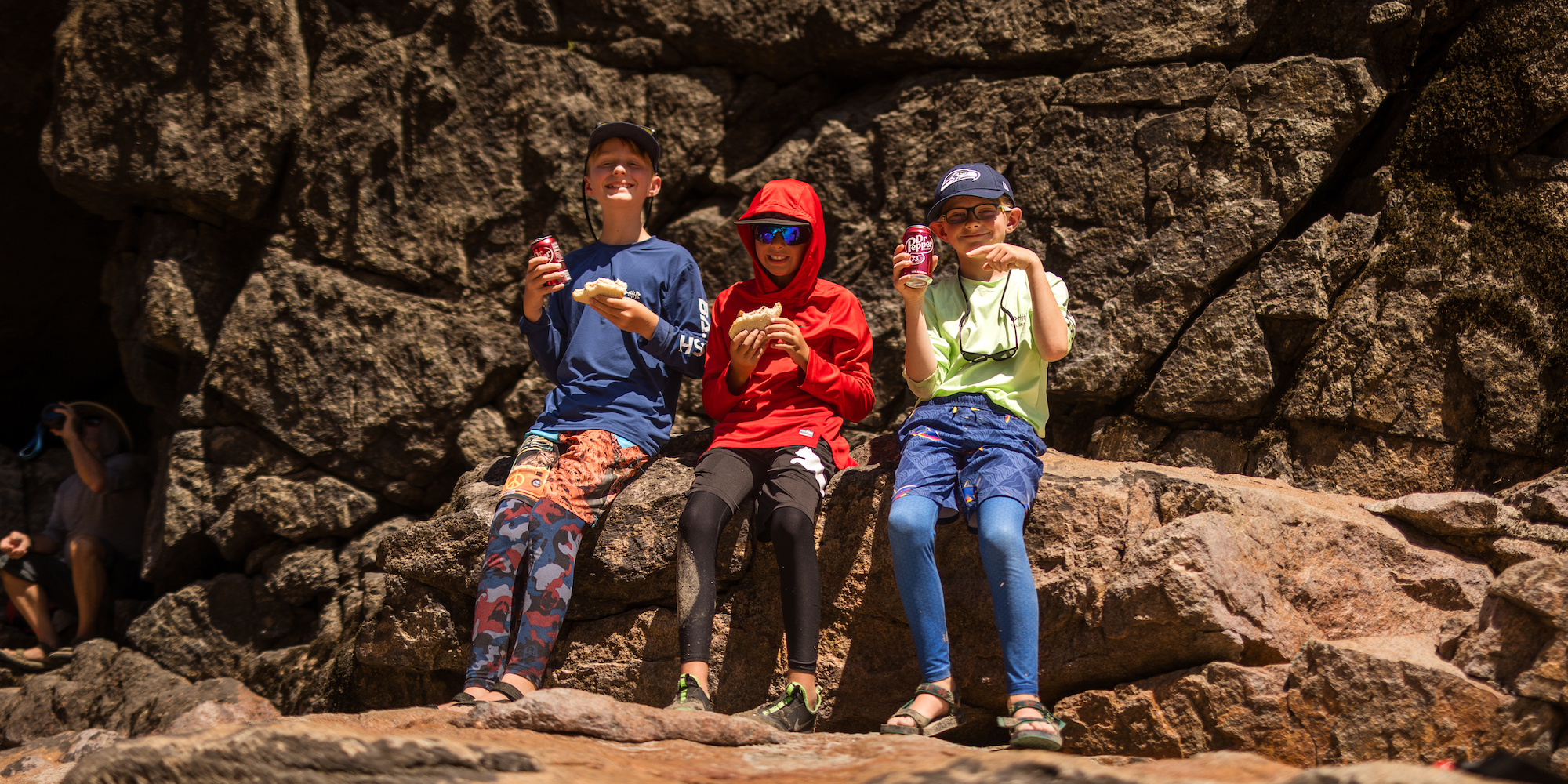 Three boys on a hike along Idaho's Salmon River holding soda and a sandwich 