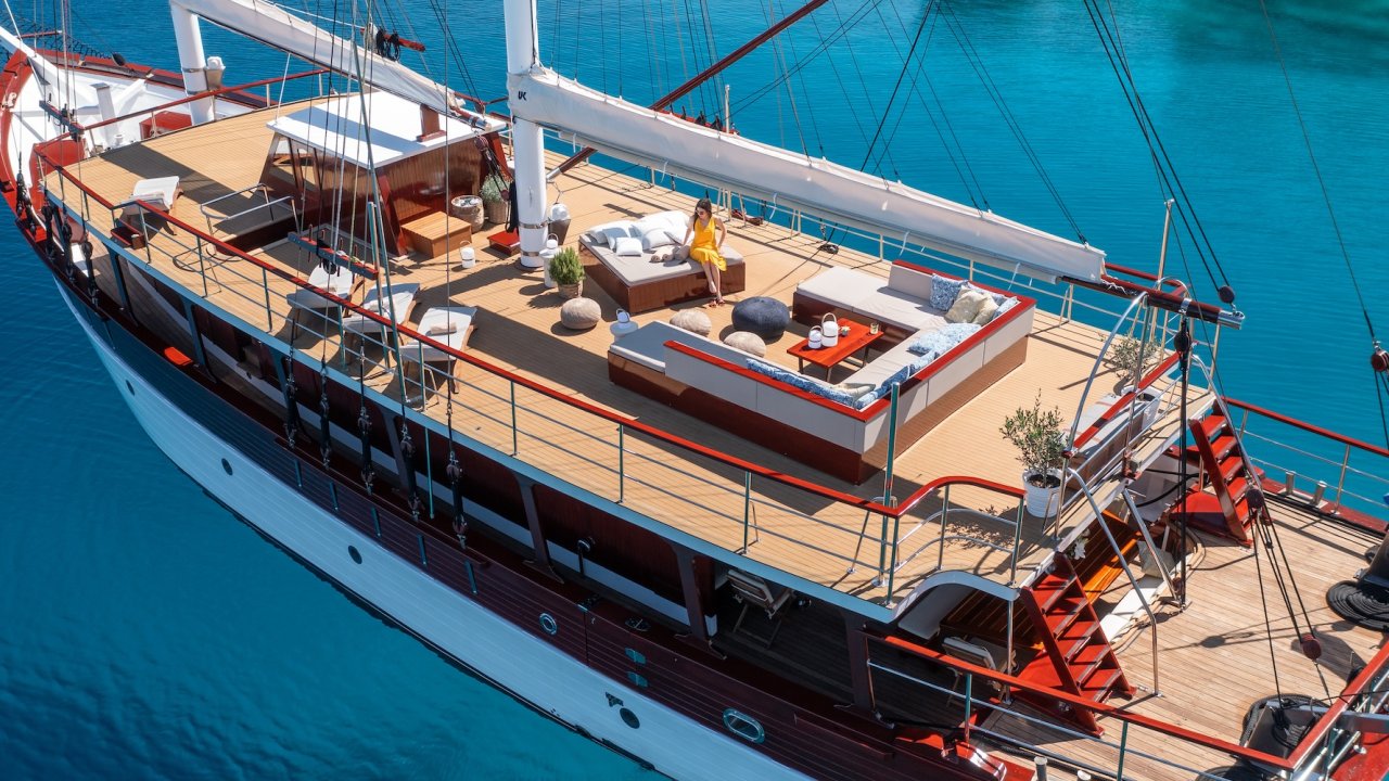 A luxury sailing yacht anchored in clear turquoise water, featuring a sun deck with lounge seating and a guest relaxing in the sun.