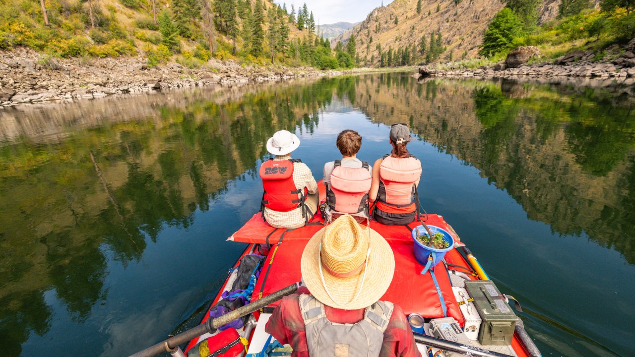 A red raft with a guide rowing and two guests sitting atop clear, still water on the Salmon River
