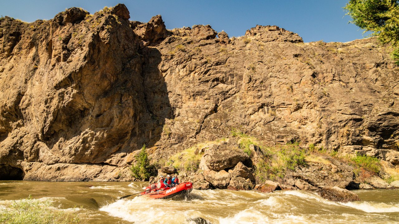 Side view of a red raft moving through a rapid with a big canyon wall behind it on a sunny day