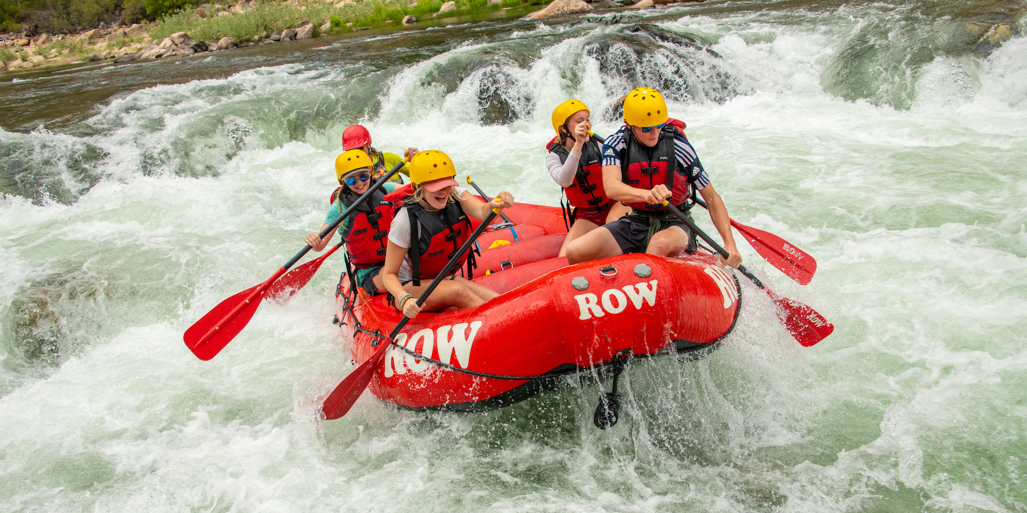 People in a red ROW raft paddling through a rapid and smiling