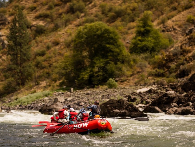A group of people in a red whitewater raft floating downstream on an Idaho river.