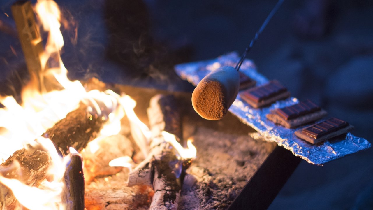 Marshmallow roasting over a campfire with s’mores ingredients nearby.