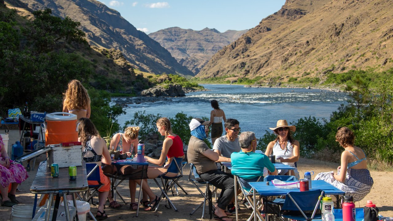 Campers sitting around square blue tables on a sandy beach along the Snake River in Idaho