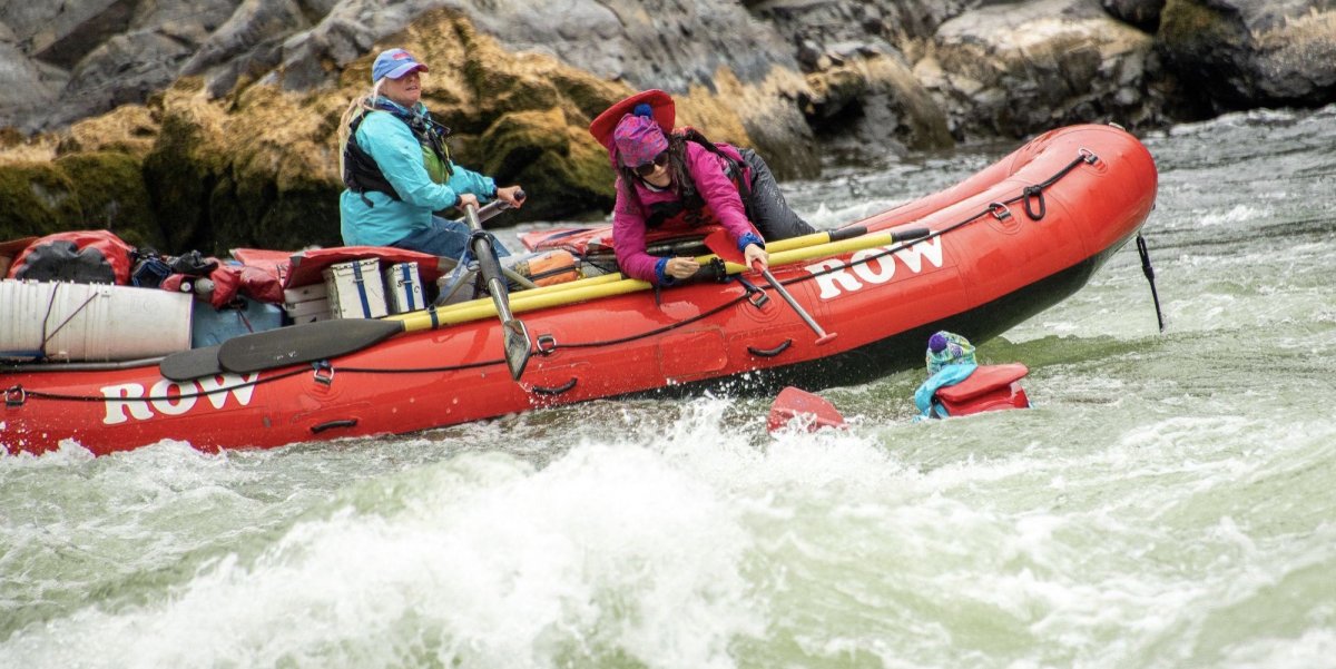 Woman on a red raft sticking her paddle out to pull a swimmer back into the boat