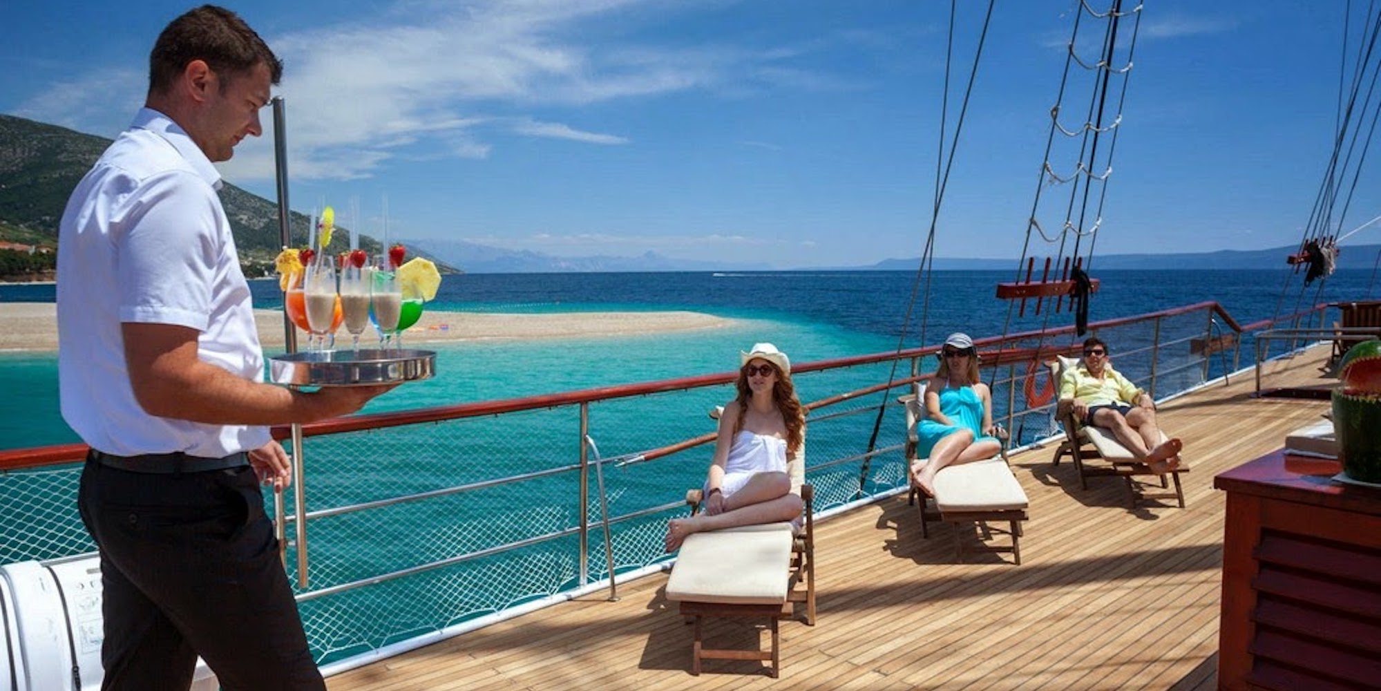 Three people sitting in lounging chairs on the sun deck of a yacht as a crew member brings them drinks