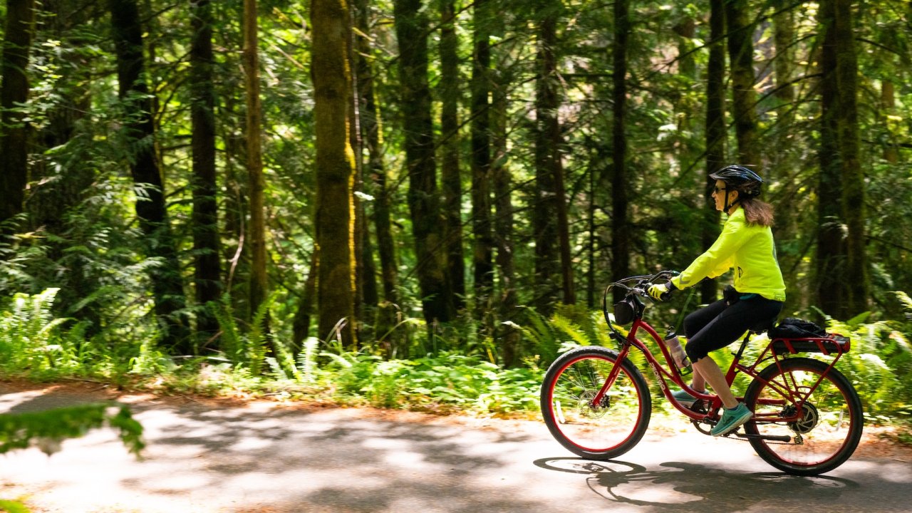 A woman in a yellow jacket rides a red e-bike through a sun-dappled forest trail in Washington State.