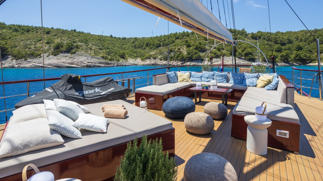 A sun deck on a yacht with cushioned longe seating, a jet ski, and views of the Adriatic Sea and green coastline.